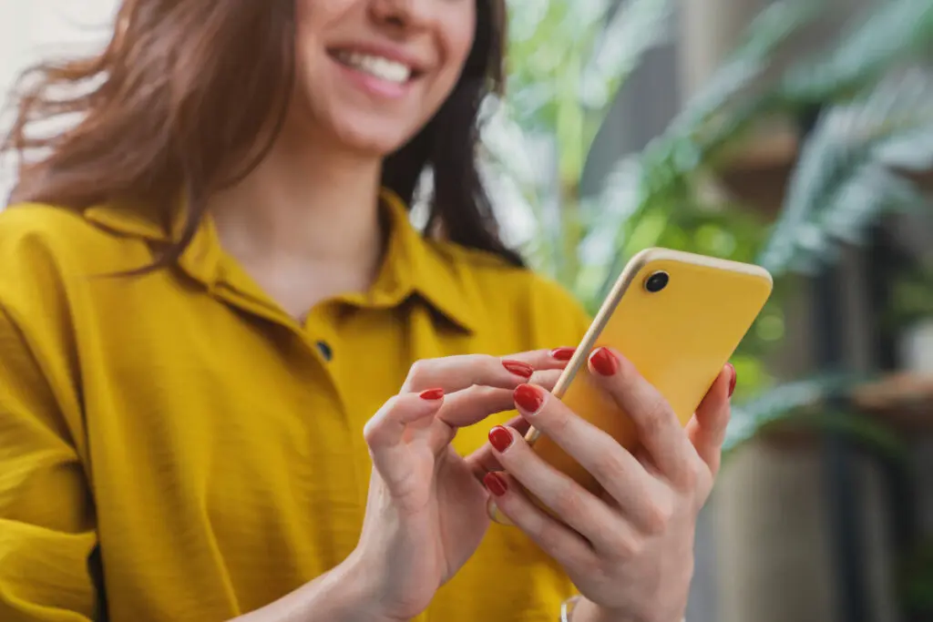 Eine Frau im gelben Shirt schaut auf ihr gelbes Smartphone und lächelt.