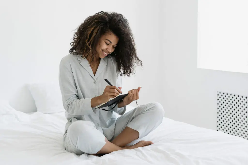 Eine Frau sitzt auf dem Bett, schaut glücklich in ihr Notizbuch und schreibt etwas auf